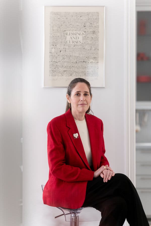 Un retrato de la Dra. Ilka Warshawsky, que viste una chaqueta roja y pantalones negros y está sentada en una pequeña silla con una obra de arte detrás de ella, que tiene las palabras Bendiciones y maldiciones legibles en el medio.