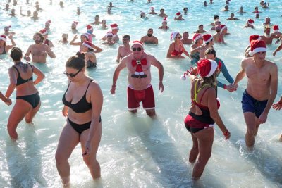 A crowd of people wearing swimsuits and Santa hats wade and swim in shallow water.